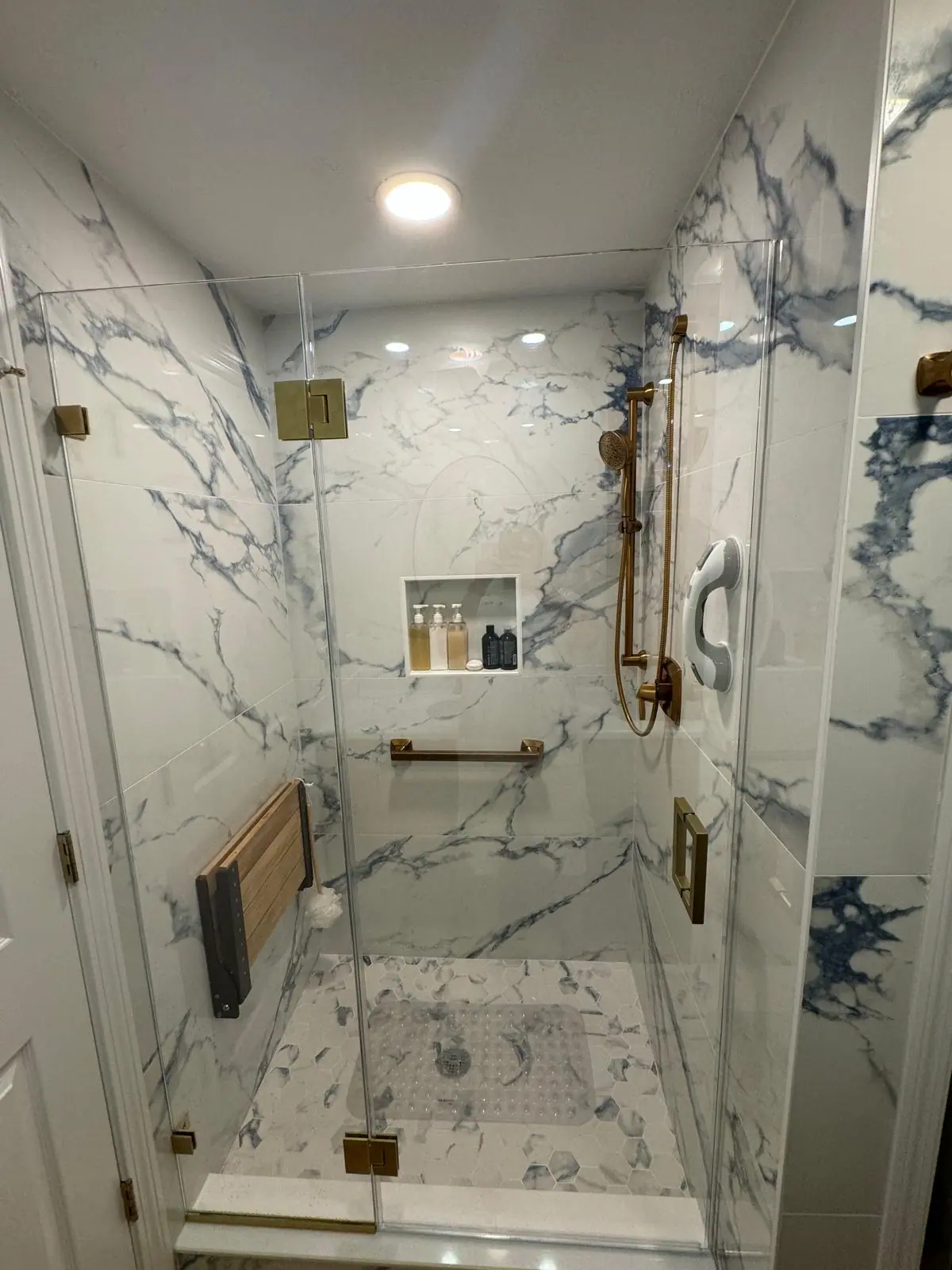 Glass walk-in shower in white porcelain with blue veining, a recessed niche, gold fixtures and a fold-down teak seat