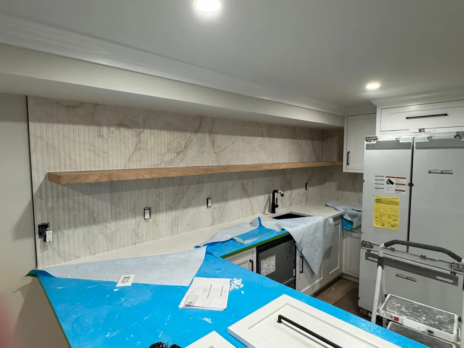 Full-height fluted marble-look tile backsplash with a floating wood shelf, countertops still protected during installation