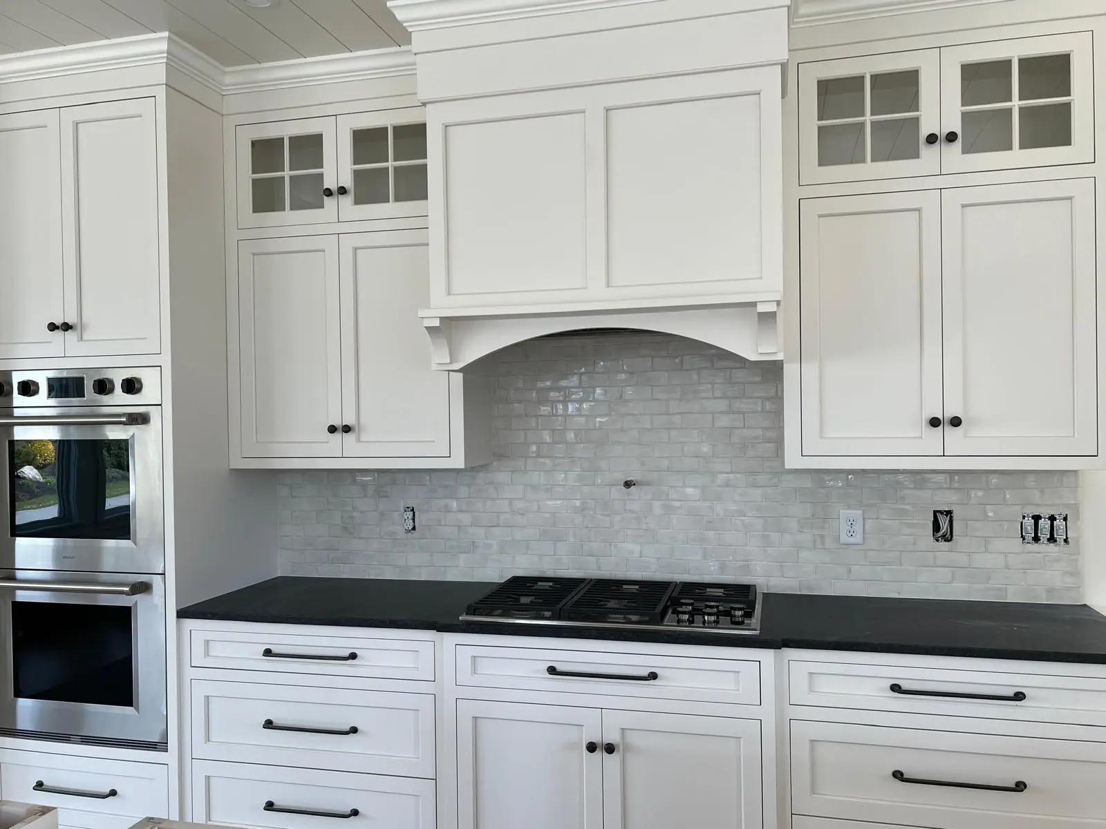 Kitchen backsplash in glossy white handmade-look subway tile under a white range hood, with white shaker cabinets and a black countertop